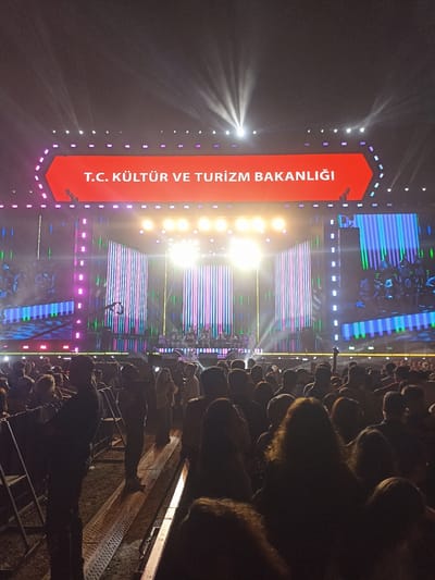 A large outdoor concert is taking place in Mezitli, Turkey. A crowd of people is gathered in front of a stage. The stage is adorned with colorful lights and vertical screens. A large red sign above the stage displays the text "T.C. KÜLTÜR VE TURİZM BAKANLIĞI." The photo is taken from the crowd. Rows of metal barriers are placed to manage the crowd. The environment suggests an evening event with stage lighting as the primary light source.