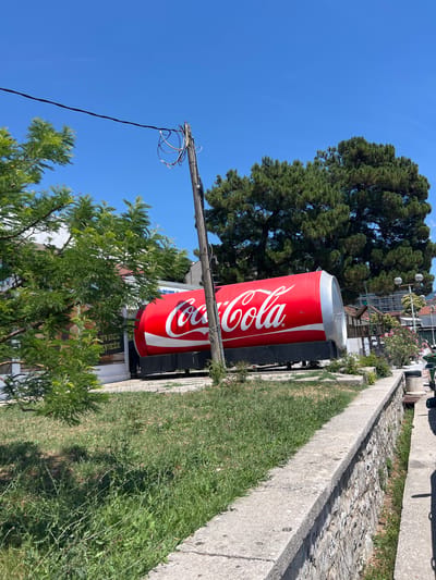 The image shows a large, red Coca-Cola can shaped structure situated outdoors in what appears to be a public space in Baošići, Montenegro. The structure is the dominant subject, with the Coca-Cola logo clearly visible in white script. It is lying horizontally, supported by a dark base. To the left, there are green trees and a building with some signage. A wooden utility pole with wires is prominent in the foreground and middle ground, to the left of the Coca-Cola can. Behind the can, a large, mature green tree provides a natural backdrop. The ground is covered with grass and some low-lying vegetation, and a concrete wall with stone construction underneath runs along the right side of the image, suggesting a pathway or elevated area. The sky is clear and blue.