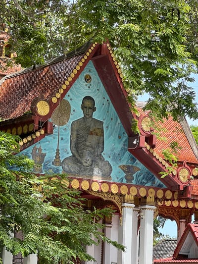This media file captures a close-up, angled view of the exterior of a building, likely a temple or shrine, in Baan Hua Thanon, Thailand. The building features traditional Thai architecture with a steeply pitched, orange-tiled roof edged with decorative gold circular motifs. The focal point is a mural painted on an angled section of the building's facade.
The mural depicts a serene, meditating figure, likely a monk, rendered in muted grey tones against a bright, turquoise blue background. The figure is seated, with hands clasped, and appears to be floating or suspended. Surrounding the figure are abstract patterns and shapes, including what appear to be stylized fish and a circular emblem with radiating lines. A string of gold circular ornaments runs along the roofline above the mural.
Lush green foliage from nearby trees encroaches on the scene, partially obscuring the building and its decorative elements. The lighting suggests it is daytime, with sunlight filtering through the leaves. The overall impression is one of cultural artistry and spiritual tranquility, set within a tropical environment. No visible text is present in the image.