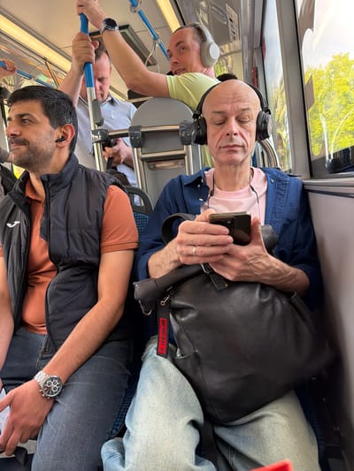 The image depicts the interior of a public transport vehicle, possibly a bus or train, with several individuals seated. The scene is set in Shcherbinka, Russia.

In the foreground, a man with dark hair and a beard is seated on the left, wearing a black vest over an orange shirt and blue jeans. He has black earbuds in his ears and a watch on his left wrist. To his right, another man, bald with headphones over his ears, is looking down at his phone. He is wearing a blue shirt over a pink t-shirt and blue jeans. A black leather bag is on his lap, with a red keychain attached.

In the background, other passengers are visible. One man in a light green shirt is holding onto a blue pole. Another man in a grey shirt is also visible behind the first man. The interior of the vehicle shows blue and grey seating, metal poles, and windows that allow natural light to enter, revealing glimpses of green trees outside. The overall impression is of a mundane commute.