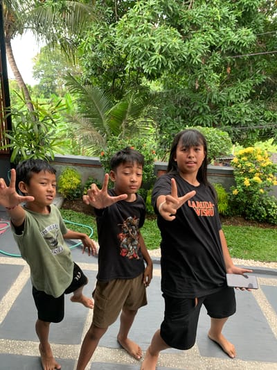 Three children pose for a photo outdoors in front of lush green foliage. On the left, a young boy in a green t-shirt and black shorts, with dark hair, extends his right hand towards the camera with his fingers spread. To his right, another young boy with dark hair wears a black t-shirt with a graphic print and brown shorts, his left hand raised with fingers spread. On the far right, a girl with long dark hair and bangs wears a black t-shirt with "GARUDA WISNU" printed in orange and black shorts. She holds her left hand up, forming a peace sign, and holds a gray smartphone in her right hand. The children are standing on a paved area with a textured surface. The background consists of dense greenery, including trees and bushes, with some flowering yellow plants visible. The lighting suggests it is daytime. The scene appears to be in a residential area with tropical vegetation, possibly in Sukasada, Indonesia.