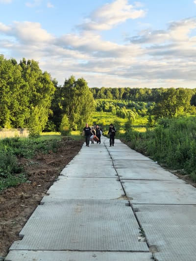 A group of five people, three men and two women, walk along a temporary concrete pathway constructed of interlocking slabs. The pathway is bordered by uncultivated land and lush green trees. The people are dressed in casual clothing. One man in the foreground carries a bag. The sky above is a mix of blue with white clouds. The scene appears to be in a rural or suburban area, possibly on the outskirts of Misaylovo, Russia, given the context. The overall impression is of a leisurely stroll or movement through an undeveloped area.