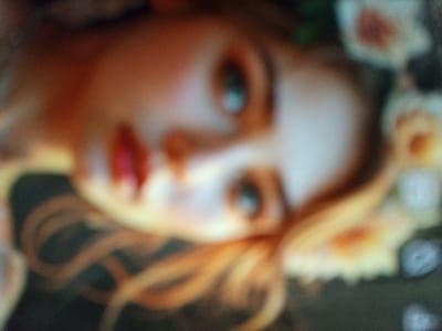 The scene is located in Kremyonki, Russia. The image displays a severely out-of-focus close-up photograph of a digital screen featuring a stylized depiction of a female subject's face. The subject's head is tilted diagonally from the upper left to the lower right. Features include fair skin, prominent light blue eyes, and red-painted lips. Golden-blonde wavy hair is visible, flowing around the face. Several white or pale yellow floral elements, possibly roses, are interspersed with green foliage along the right side of the subject's head. The background is predominantly dark, suggesting a digital interface, with faint, indistinguishable graphical elements visible along the far right edge of the frame. No discernible actions or interactions are present within the static depiction.