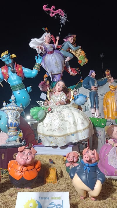This image captures a large, multi-component artistic monument, consistent with a "Falla" structure, displayed outdoors at night in El Port De Sagunt, Spain. The scene features numerous anthropomorphic figures set against a dark background with minimal ambient lighting.
At the upper level, a white-haired woman in a purple and gold gown, holding a staff with a starburst and pink swirling ornament, appears to float or dance. Adjacent to her, a man in a teal tunic, pink hat, and striped pants, is depicted in a dynamic leaping posture, carrying a cornucopia overflowing with colorful fruits including pumpkins, watermelons, grapes, and apples.
The mid-level prominently features a large, blue Genie-like figure with a red vest and gold accessories on the left, gesturing with an open palm. To the right of the Genie, a woman in an elaborate white and gold dress is seated on a golden armchair, holding a small green fairy-like figure that holds a red apple. Behind her, a man in a dark blue outfit and purple head covering stands, holding a lantern-like object. A woman in a yellow dress is partially visible beside him. Smaller figures, including a floating pink-dressed doll and a blue-dressed doll, are positioned around the seated woman.
The lower section of the monument, serving as its base, includes an ornate blue and white urn-like structure on the left. The ground plane is covered with hay or straw. Three anthropomorphic pig figures are visible on this level: one on the left in an orange jacket and black pants, one on the right in a blue jacket, blue scarf, and tan pants, and a smaller, partially obscured pig in a green garment between them. A small, framed plaque with text is positioned on the straw. In the immediate foreground, a white sign with a blue sun logo and the text "Escuela Infantil Sol" is visible.