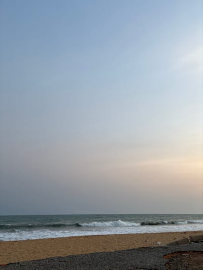 The photograph depicts a coastline view, with the sky occupying the majority of the frame. Its gradient transitions from a soft blue to a pale yellow-grey towards the top. A narrow strip on the right reveals a beach where gentle waves meet the shore, the sand adjacent to a dark, rocky boundary. The overall atmosphere is serene, with a hazy quality obscuring finer details. The photograph's orientation is vertical.