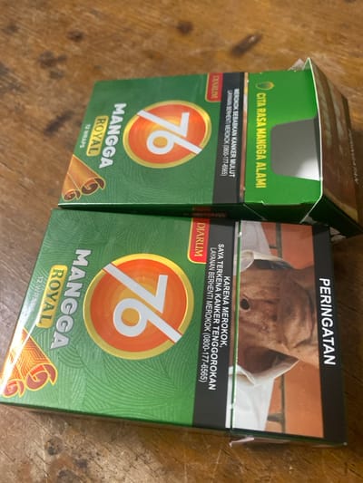 The image displays two packs of Mangga Royal 76 kretek cigarettes by Djarum, oriented diagonally on a brown wooden surface. Both packs are predominantly green with a central orange and gold circle containing the number "76" and the text "MANGGA ROYAL". Each pack indicates "12 WRAPS".
The upper pack is partially open at the top, revealing a grey interior flap. Its visible side features a black band with Indonesian text: "DIARUM" and "MEROKOK SEBABKAN KANKER MULUT LAYANAN BERHENTI MEROKOK (0800-177-6565)" (Smoking causes oral cancer. Quit smoking service (0800-177-6565)). Adjacent to this is green text stating "CITA RASA MANGGA ALAMI" (Natural Mango Flavor).
The lower pack is closed and presents a graphic health warning on its side. This warning shows a close-up image of a person with a visible surgical stoma on their throat. Accompanying Indonesian text reads: "PERINGATAN" (WARNING) vertically, and horizontally, "DIARUM", "KARENA MEROKOK, SAYA TERKENA KANKER TENGGOROKAN LAYANAN BERHENTI MEROKOK (0800-177-6565)" (Because of smoking, I have throat cancer. Quit smoking service (0800-177-6565)).
No individuals are present in the scene. The wooden surface exhibits a distinct grain pattern. The scene is located in Cimenyan, Indonesia.