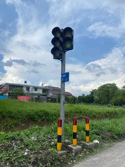 The image shows a traffic light with a green light, located on a street in Margaasih, Indonesia, at coordinates -6.97 latitude and 107.551 longitude. The traffic light is mounted on a gray metal pole, with three yellow and black striped posts positioned in front of it. A blue sign above the light says "K Kiri Terus," which translates to "Left Turn Straight." The pole is surrounded by lush green grass and bushes. In the background, a building with a red sign that says "KRIJ TRIO KERAMIK & GRANIT KUALITAS 1" can be seen. The sky is blue with white clouds.