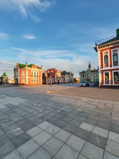 A wide-angle shot captures a European-style town square paved with grey stone tiles. The late afternoon sun casts long shadows across the scene, illuminating the ornate architecture of the surrounding buildings. These buildings feature vibrant red and green facades with intricate white detailing, arched windows, and steeply pitched green roofs adorned with spires.

On the right, a modern brick building with white trim and arched windows dominates the foreground. To its left, a row of similarly styled buildings continues, creating a cohesive and picturesque streetscape. A silver car is parked in front of a striking green building with a red-topped spire in the distance. A single person walks on the sidewalk in front of the orange buildings, while another is seen in the far background.

The sky above is a brilliant blue, dotted with wispy white clouds, suggesting fair weather. The overall atmosphere is tranquil and bathed in the warm glow of the setting sun. The location appears to be a historic or well-preserved urban center, likely Yoshkar-Ola, Russia, given the architectural style. No visible text is discernible in the image.