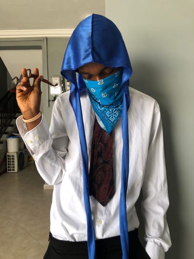 A dark-skinned individual stands indoors in Gbawe, Ghana, wearing a blue satin durag covering the head and a light blue bandana with a white paisley pattern obscuring the lower face. The person is dressed in a white long-sleeved collared shirt, black trousers, and a dark tie featuring a red and black paisley pattern. A light-colored wristband is present on the left wrist. The individual's gaze is directed downward, and the left hand holds a pair of brown-framed eyeglasses by one temple piece. The background consists of a light grey wall. To the left, a portion of an indoor staircase with white risers is visible, along with a white electrical meter mounted on the wall. The floor is covered with light-colored tiles, and two white cylindrical buckets are stacked next to a white rectangular air conditioning unit on the far left.