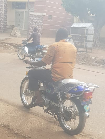 In Kufena, Nigeria, a man in a mustard-colored shirt and dark pants is seated on a motorcycle on an unpaved road. Another man, wearing a brown and black patterned shirt and dark pants, rides a similar motorcycle in the background. Buildings and trees line the street.