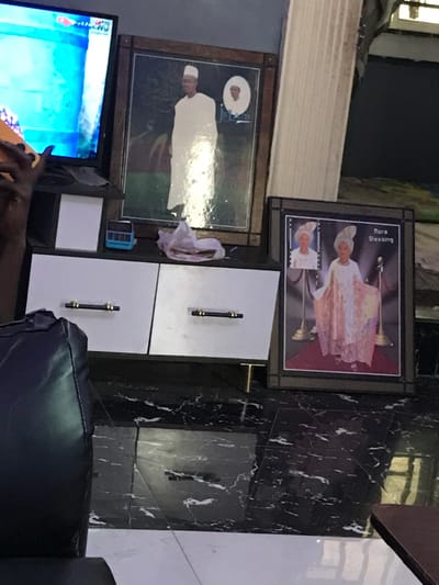 Two framed pictures are on a black and white marble wall above a white cabinet with two drawers. The top picture shows a person in an ornate outfit. The picture below shows a person in white attire. A small blue digital device sits on a shelf to the left of the cabinet. To the left is a television displaying a bright, colorful scene. A dark leather chair sits in the foreground.