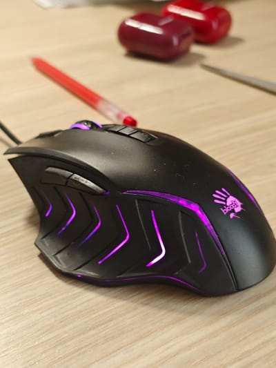 A black gaming mouse with purple LED lighting sits on a light-colored wooden desk. The mouse has a distinctive ergonomic design with angled lines and a textured grip area. The "Bloody" logo, a stylized handprint, is visible on the right side of the mouse. In the background, out of focus, are two red objects that appear to be caps or small containers, and a red pen with a transparent casing. A thin, metallic object, possibly a file or a thin tool, is also present in the background, to the right of the red objects. The lighting suggests an indoor setting, and the overall impression is that of a workspace or a gaming setup. There is no indication of the time of day or weather conditions. The location context is Svietlahorsk, Belarus.