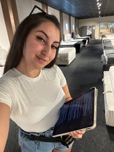 The media file shows a young woman, likely in her late teens or early twenties, taking a selfie in what appears to be a mattress store. She is holding a tablet in her right hand, displaying text, and looking towards the camera with a slight smile. She is wearing a white t-shirt adorned with small, sparkling embellishments and blue denim shorts. Her dark hair is down, and she has glasses perched on top of her head.
The background reveals rows of mattresses and bedding products, suggesting the setting is a retail environment. The lighting appears to be artificial, typical of an indoor store. The overall mood seems casual and relaxed, possibly indicating the woman is browsing or on a break.
The tablet screen shows text, which is too small to be clearly deciphered, but the interface suggests a reading or note-taking application. The location context provided is Targovishte, Bulgaria, which adds a geographical element to the scene. There are no clear indicators of the time of day or weather conditions, as the scene is indoors.