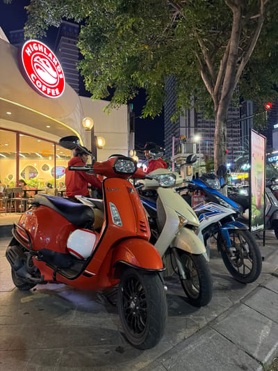 A nighttime scene in South Quy Nhon Ward, Vietnam, features several parked scooters and motorcycles lined up along a sidewalk. The foreground is dominated by a bright orange Vespa scooter, with a white helmet adorned with red and green stripes resting on its seat. Behind it, a beige scooter and a blue and white motorcycle are also parked.
The scooters are positioned in front of a building with a large, illuminated sign that reads "HIGHLAND COFFEE" in a red circular logo. The building's facade has large windows, revealing a warmly lit interior with patrons inside. Outdoor lighting fixtures, resembling spherical lamps, hang above the coffee shop entrance, casting a soft glow on the scene.
A large tree with leafy branches extends from the left side of the frame, its trunk and limbs providing a natural element to the urban setting. In the background, tall buildings and streetlights indicate a bustling city environment. The overall impression is one of a common urban scene at night, likely a busy area with a popular coffee shop. The image is captured in portrait orientation.
