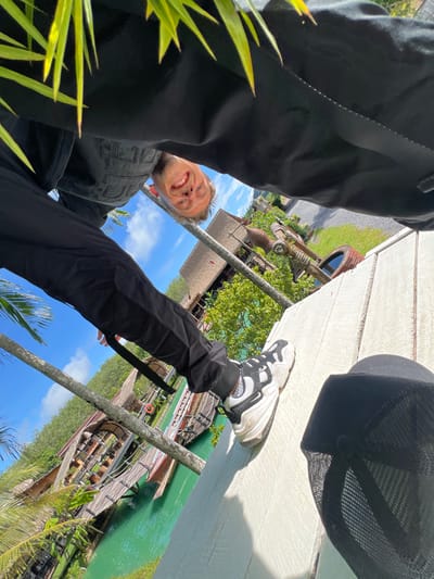 This is an upside-down, low-angle selfie taken outdoors. The main subject is a young man with blonde hair, blue eyes, and a wide smile, looking directly at the camera. He is wearing black pants and a black denim vest over a black shirt. His left leg is bent, with a white and black athletic shoe visible. A black baseball cap with a mesh back is lying on a light-colored wooden surface in the foreground.
The background features a vibrant green body of water with wooden walkways and small boats. Several thatched-roof structures are visible along the water's edge, surrounded by lush green trees and palm trees. In the distance, there are more trees and what appears to be a road. The sky is bright blue with scattered white clouds, suggesting a sunny day.
The scene is set in a tropical or subtropical location, with the water, vegetation, and architectural style hinting at a resort or a natural park. The overall mood is cheerful and relaxed, conveyed by the man's enthusiastic smile. The time of day appears to be daytime, likely late morning or early afternoon due to the bright sunlight and clear sky. No text is visible in the image. The location context provided is Tambon Pa Kae Bo Hin, Thailand.