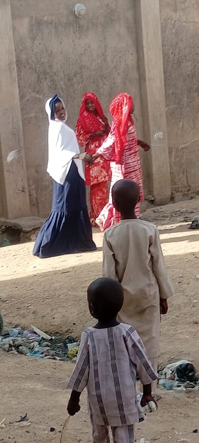 The image depicts an outdoor scene in Rijiyar Zaki, Nigeria, featuring five individuals under clear daylight. In the midground, three women are gathered near a rough, light-colored wall. The woman on the left wears a white hijab and a dark blue skirt, smiling directly at the camera while holding hands with another woman dressed in a vibrant red patterned garment and headscarf. A third woman, similarly attired in red, stands to their right, partially obscured.

In the foreground, two young children are seen from behind, walking or standing on unpaved, sandy ground. The taller child wears a light beige tunic. The smaller child in front wears a light-colored, vertically striped tunic and trousers, holding a thin stick in his right hand and a small white object in his left. Litter and debris are scattered on the ground to the left.

The setting appears to be a casual street or open area, with the wall showing signs of wear. Faint markings, including the numbers "1907", are visible on the wall in the background. The interaction among the women, particularly the smiling face of the woman in white, suggests a friendly or joyful encounter.