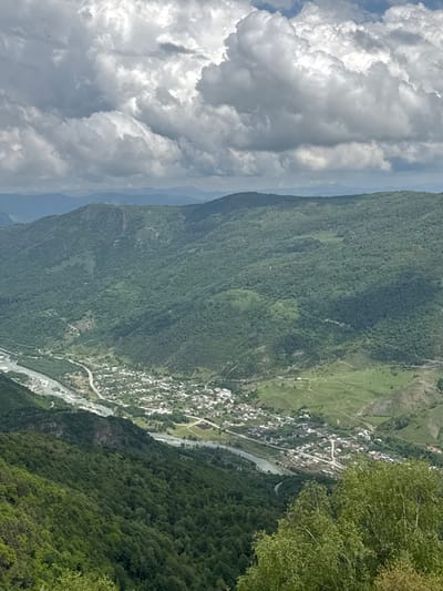 The image captures a panoramic view of a valley with a village nestled within it, bordered by a river and flanked by densely forested mountains. The sky is filled with dramatic, heavy clouds, suggesting an overcast day. In the foreground, lush green trees, likely birch trees, dominate the bottom right of the frame, with their light green foliage reaching upwards. The middle ground reveals the village of Nizhnyaya Teberda, Russia, characterized by numerous small houses with red and gray roofs, spread out along a road that follows the winding path of a river. The river itself is wide and appears to have a rocky bed, with its water reflecting the muted light of the sky. The background consists of rolling, verdant mountains covered in thick forests, their slopes crisscrossed with visible roads or paths. In the distance, further mountain ranges are visible, their forms softened by atmospheric haze. The overall scene conveys a sense of remote, natural beauty and a settled, rural community within a dramatic mountainous landscape.