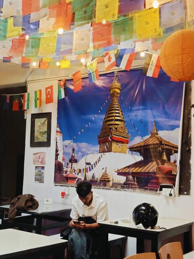 A man sits at a table in a restaurant, looking at his phone. He is wearing a white shirt and blue jeans. A black helmet sits on the table next to him. The restaurant walls are decorated with prayer flags and a large mural of a stupa. The prayer flags are colorful and strung across the ceiling. The mural depicts a golden stupa with Buddhist eyes, set against a blue sky with clouds. A brown jacket is draped over a chair at another table. There are also several small framed pictures and posters on the wall. The lighting is dim, with some small bulbs illuminating the prayer flags. The overall impression is that of a casual eatery, possibly catering to tourists.