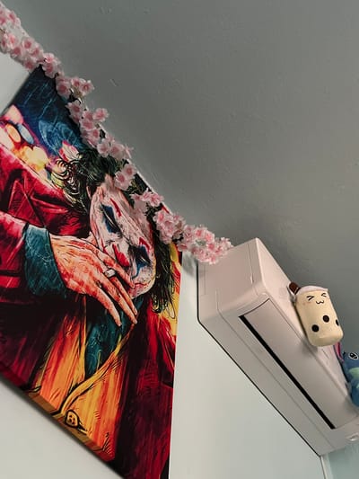 A low-angle medium shot captures a portion of a wall, a white air conditioning unit, and a canvas print of the Joker character. The canvas, which takes up the left two-thirds of the frame, depicts the Joker with his hands clasped near his face, his white and red makeup prominent. A garland of small pink flowers drapes across the top edge of the canvas and extends slightly towards the air conditioner. The air conditioner is positioned on the right side of the frame, mounted on the wall above the canvas. Perched on top of the air conditioner is a plush toy resembling a cup of bubble tea, adorned with a smiling face. Partially visible on the right edge of the air conditioner is another plush toy, blue and with black eyes. The wall is painted a light blue, and the ceiling is visible at the top of the frame. The scene appears to be an indoor domestic setting. There are no visible geographical indicators that would suggest a location in Debelt, Bulgaria.