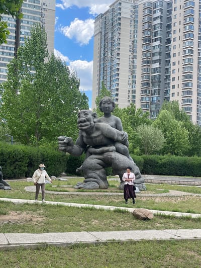 Here's a matter-of-fact description of the image:

The image shows a park scene in Changping District, China, featuring a large grey statue of a child on top of an animal-like figure. A tall apartment building is visible in the background under a blue sky with puffy white clouds. Two people are in the frame: an older woman in a light-colored jacket and dark skirt standing next to the statue and a person wearing a hat, light-colored shirt, and dark pants walking along a stone-lined path. The ground is covered with grass.