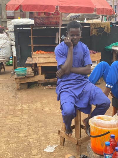 The image captures a man in Kafanchan B, Nigeria, engaged in a phone conversation while seated on a wooden structure, possibly a makeshift bench or stall. He is dressed in a blue, flowing garment. The scene is an outdoor market environment with stalls selling various goods. To the man's right is a wooden platform holding a covered orange bucket and several bottles of what appears to be a bottled beverage. In the background are stalls and various produce, including tomatoes. Underneath, in front, and behind the stalls are bags, tarps, various buckets, and waste debris that is found in open-air market settings. Overhead are red awnings. The ground is patterned with what appears to be concrete. A sign for the Kafanchan Modern Market is partially visible. Another person is seated above the man in a similar-style blue garment with white-striped pants.