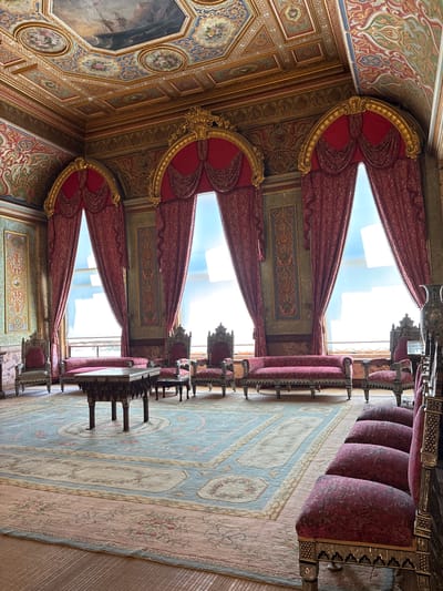 This image depicts a richly decorated interior, likely a palace or grand hall, in Beşiktaş, Turkey. The scene is characterized by opulent furnishings and intricate architectural details. Dominating the foreground and mid-ground are several rows of plush, upholstered seating, upholstered in a deep red, patterned fabric. These seats appear to be part of elaborate benches and armchairs with ornate, dark wooden frames, possibly inlaid with decorative elements.

The ceiling is a focal point, adorned with detailed frescoes and gilded moldings. Large, arched red draperies frame tall windows that reveal a bright, likely daytime sky. The windows themselves are tall and narrow, suggesting a historic architectural style.

A large, intricately patterned rug covers the floor, featuring a palette of muted blues, creams, and pinks, with floral motifs. Several ornate tables, with dark, detailed bases, are positioned between the seating arrangements. The walls also feature decorative panels and colorful, patterned artwork.

The overall impression is one of grandeur and historical significance, suggesting a formal or ceremonial space. The lighting suggests it is daytime, with sunlight filtering through the windows. No people are visible in the image, and there is no visible text. The scene evokes a sense of historical elegance and luxury.