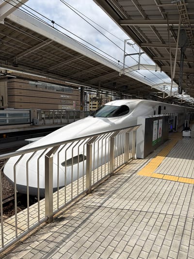 The image depicts a Shinkansen bullet train stopped at a station in Kyoto, Japan. The train, primarily white with a sleek, aerodynamic design, occupies the left side of the frame, its front section prominently visible. It is positioned on a track adjacent to a platform. The platform is paved with light-colored rectangular tiles and features a yellow tactile paving strip near the edge. A silver metal railing with vertical bars separates the platform from the train tracks.

In the background, a multi-story building with horizontal wooden paneling is visible. Signage on this building reads "DRUM TAO" and "KYOTO AVANTI," suggesting a commercial or entertainment complex. Above the train and platform, a covered structure with a metallic roof supported by beams is present, with overhead electrical wires for the train's power. The sky above is partly cloudy.

To the right of the train, a silver rolling suitcase stands alone on the platform. A few advertising posters are affixed to a vertical structure near the train doors. The scene is well-lit by natural daylight, casting shadows from the railing and overhead structure onto the platform tiles. No people are visible in the image. The overall environment suggests a functional, modern transportation hub.