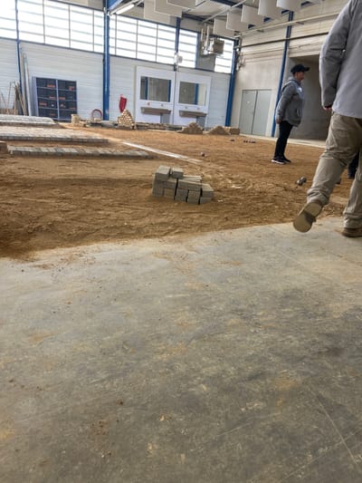 The image captures an indoor pétanque court within a large, high-ceilinged industrial-style building in Montigny-lès-Metz, France. The foreground consists of a concrete floor, transitioning into a large expanse of packed earth and sand that forms the playing surface. The building's walls are constructed from white corrugated metal with visible blue vertical support beams and horizontal window sections at a higher elevation. White acoustic panels are suspended from the ceiling.
On the left side of the playing area, stacked rows of grey rectangular pavers are arranged, suggesting boundary markers or targets. Several loose stacks of identical grey pavers are distributed across the sandy ground, with a prominent stack of approximately 16 blocks positioned in the mid-ground. Multiple metallic pétanque balls are visible on the court, including individual balls and a cluster of three further back.
Two individuals are present: a woman stands in the mid-ground, facing left and observing the court. She wears a grey hooded sweatshirt, dark pants, and a dark baseball cap. In the immediate right foreground, the lower half of another person is visible, wearing light brown trousers and light-colored shoes, stepping away from the camera. The legs of a third person are partially discernible in the background, further to the right. The back wall features two recessed white structures with shelves below, a double door to the right, and a blue metal storage cabinet on the left. Construction materials are visible leaning against the wall on the far left. A red cylindrical object, possibly a fire extinguisher, is mounted on the back wall near the center.