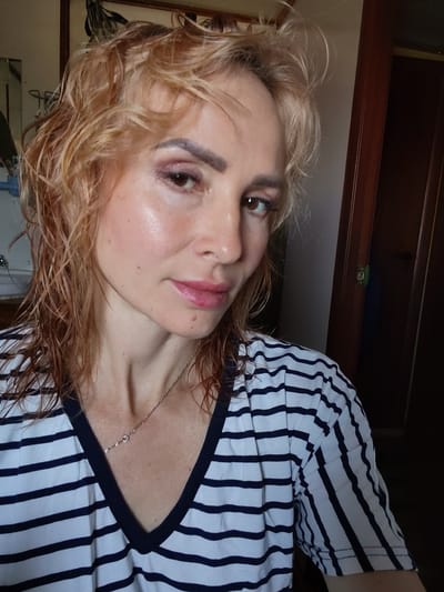 A woman with blonde, wavy hair and fair skin is pictured in a close-up shot, looking slightly to her left. She is wearing a striped t-shirt with navy blue and white horizontal stripes, and a thin silver necklace. Her makeup includes eyeshadow and lipstick. The background is dimly lit, with a wooden door visible on the right side of the frame. The setting appears to be indoors, possibly a room in a residential building, and there is no indication of it being in Kvartsitny, Russia, or any specific city.