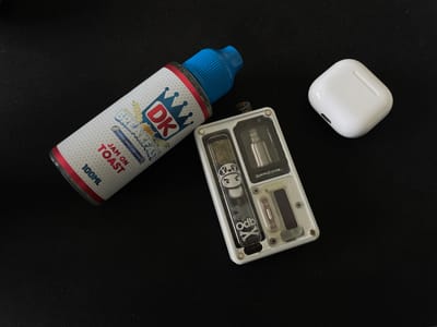 An overhead shot shows a black surface with three electronic items arranged in a triangular formation. On the left, a white bottle with a blue cap and a label featuring a crown and "DK BREAKFAST" is angled diagonally. The label also reads "JAM ON TOAST" and "100ML". In the center, a white rectangular vaping device with a clear front panel is positioned vertically. Visible through the panel are metallic components and decorative elements, including a cartoon character and the text "Odb X". To the right, a white, square wireless earbud charging case is placed. The environment appears to be indoors, with a dark, textured surface that could be a desk or cloth mat. There are no people or visible signs of activity. The image does not contain any information about the city of Voskresensk, Russia.