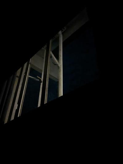 The media file is a photograph taken at night in General Trias, Philippines. It features a dark, abstract composition with a section of wooden framing illuminated by a light source that is not visible in the frame. The framing appears to be part of a window or opening, with vertical and diagonal wooden beams creating a geometric pattern. A dark, reflective surface, possibly glass, is visible within the frame, hinting at the presence of a window. A small, yellow object, possibly a latch or handle, is attached to one of the beams. The background is entirely black, suggesting a very dark environment or the absence of any discernible background elements. The overall impression is one of an unfinished or partially constructed structure seen in low light conditions. There is no text visible in the image.