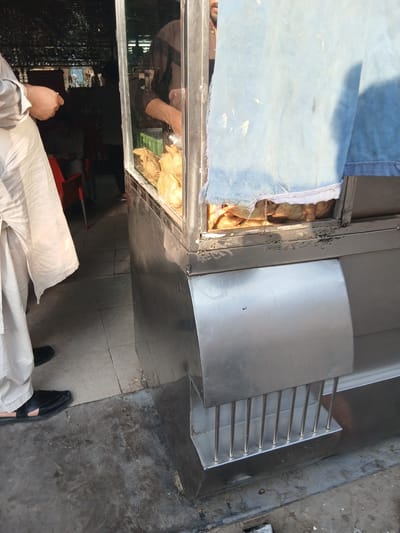 The image depicts an outdoor food stall in Peshawar City Tehsil, Pakistan. A person wearing a light-colored shalwar kameez and black sandals is visible on the left, facing away from the camera. To the right, a stainless steel food display case contains several fried food items, possibly samosas, illuminated from within. A blue cloth is draped over the top of the stall. The ground is a mix of concrete and tiled flooring. In the background, hints of a dimly lit interior with seating and other people suggest a bustling environment. The stall appears to be situated in a market or street setting.