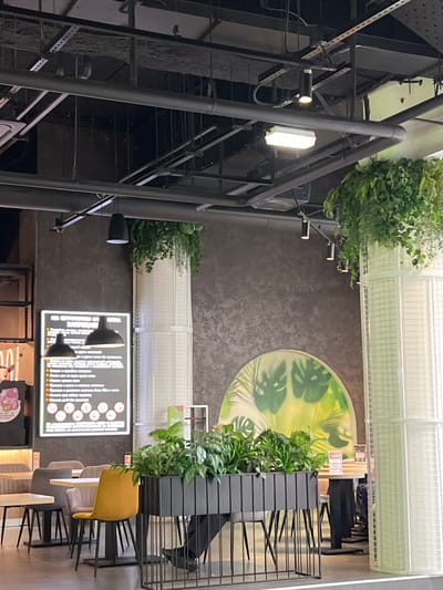 The image depicts an indoor setting, possibly a restaurant or cafe, with a modern and industrial aesthetic. A prominent feature is a large, dark metal structure with multiple levels and railings, resembling scaffolding or a multi-story shelving unit. Lush green plants are strategically placed throughout the space, some hanging from above and others in planters on shelves, adding a touch of nature.

In the background, a circular mural adorns a dark textured wall, featuring stylized green leaves and tropical foliage against a light yellow background. Further back, a white structure with a grid pattern spans the ceiling, possibly a skylight or architectural element.

In the foreground, a black menu board is displayed, illuminated by two cone-shaped black lights. The menu itself contains white text and circular icons, though the content is illegible. Several chairs are visible around tables. One chair is a bright yellow, while others are grey and white. A black metal shelving unit with a slatted design is positioned next to the chairs.

The lighting appears to be artificial, with a mix of warm and cool tones. The overall impression is a contemporary space that combines industrial design with natural elements. No people are clearly visible in the image, and the activity appears to be static, as if captured during a moment of pause. The image is oriented vertically.
