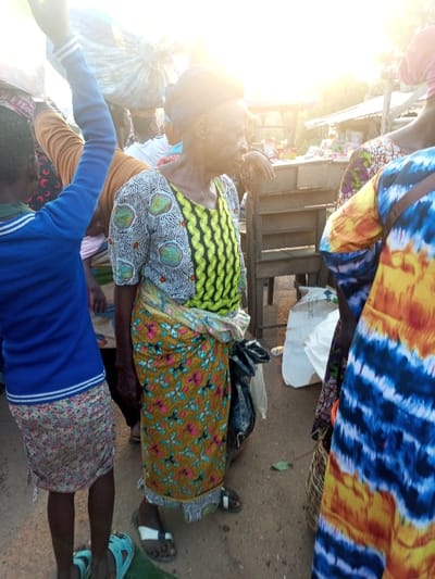 An outdoor scene captures several individuals, predominantly women. An elderly woman with distinct facial lines stands out, wearing a patterned top with a yellow and black panel and a colorful skirt. To her left, a child in a blue top raises an arm. Other figures surround them, some with goods balanced on their heads or wearing brightly colored clothing. A market stall is partially visible in the background, adding to the busy atmosphere. The sunlight is bright, and the ground appears to be a dirt road.