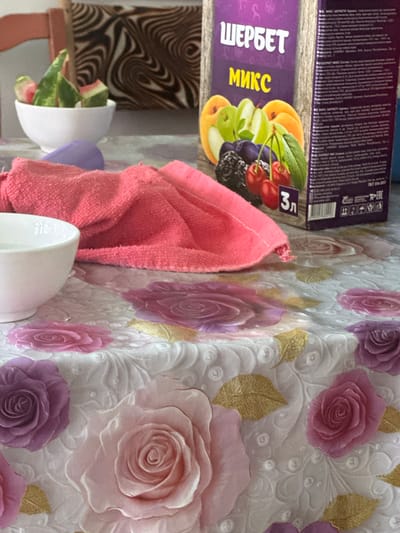 The image depicts a close-up view of a table with a floral-patterned tablecloth. On the table, a white bowl filled with watermelon rinds is visible. Next to it, a pink towel is draped. A carton of juice with Russian text and fruit imagery, specifically cherries, plums, apples, and apricots, stands to the right. The background shows a portion of a chair with a zebra-like pattern. The scene appears to be an indoor setting, possibly a home or a cafe. There is no specific indication of the city of Тайгараев, Kyrgyzstan, in the image.