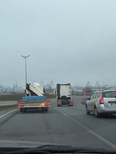 The image depicts a daytime scene on an overcast day in Agigea, Romania, showing a section of a multi-lane road transitioning towards a port area. The road surface is asphalt, with visible tire marks in the lane closest to the viewer. In the midground, a white Volvo truck cab with a folded white truck-mounted crane is positioned on a blue flatbed trailer, displaying the Romanian license plate SB 99 ARA and "CONVOI EXCEPTIONNEL" text. Its rear amber marker lights and red tail lights are illuminated. To its right, in an adjacent lane, is another white Volvo truck cab (tractor unit only), with its amber turn signals or hazard lights active. Further to the right, a silver Dacia Logan MCV station wagon, featuring green, orange, and red graphics on its side, is partially visible, displaying a Romanian license plate starting with IS 17. Its red tail lights are also active.

In the background, beyond a grassy median and a traffic roundabout indicated by a red-bordered white Yield sign and yellow chevron signs, lies an extensive industrial port complex. Numerous large gantry cranes and other port machinery are visible along the waterfront, with several cylindrical storage silos situated closer to the road. A tall, gray street light pole with a dual-arm fixture stands on the left side of the road. The sky is uniformly light gray, indicative of heavy cloud cover. No individuals are explicitly visible in the scene.
