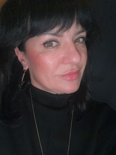 A woman with black hair and fair skin is lying down, her head tilted to the side. She has green eyes, pink lipstick, and blush on her cheeks. She's wearing a black turtleneck sweater and a thin gold necklace. An earring is visible on her left ear. The background is dark and blurry. The image appears to be taken at a close-up angle.