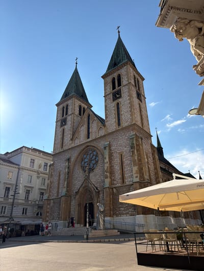 The image displays a low-angle view of the Sacred Heart Cathedral in Sarajevo, Bosnia and Herzegovina. The cathedral, with its twin spires and Gothic architectural style, dominates the frame. The stone facade is detailed with arched windows and clock faces visible on one of the towers. A large rose window is present on the main building. Sunlight illuminates the scene, suggesting it's a clear day, possibly late morning or early afternoon, with a bright blue sky and scattered clouds.
In the foreground and to the right, a street cafe is set up with tables and chairs under a large yellow umbrella. People are seated and standing at the cafe, enjoying the outdoor space. Further to the right, people are also seen walking on the paved street and sidewalk. To the left and at the top of the image, architectural details of a building, possibly a neighboring structure or part of the cathedral's surroundings, are visible, including a sculpted figure. The overall atmosphere appears pleasant and serene, with people engaging in everyday activities in a historic urban setting. Text is visible on the umbrella, reading "Coca-Cola".