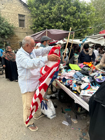The image depicts an outdoor scene with several people amidst a cluttered stall filled with various items, including clothing, toys, and bags. The stall appears to be set up along a street, with a building made of stone in the background.
In the foreground, a man in a striped shirt and khaki pants is holding an American flag. His hands are visible as he grasps the flag, which is draped over his legs. He is wearing sandals. To his left, a woman in a black abaya and patterned hijab is visible.
Behind the man holding the flag, other individuals are present, some looking towards the stall's contents. The stall is overflowing with goods, suggesting a marketplace or a sale. There's a glimpse of a bicycle or motorcycle in the lower left corner.
The lighting suggests it might be daytime, possibly overcast given the diffused light. The overall atmosphere seems to be that of a busy, informal gathering or trading area. There is some visible text on a white bag in the mid-ground, partially obscured, but it appears to contain letters like "N" and "AG". A white book with "Disney Solo's For Kids" is also visible among the merchandise. The setting appears to be a street in Karachi Division, Pakistan, given the provided location context.