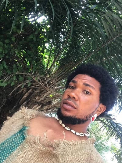 A low-angle portrait depicts a single individual, appearing male, with dark curly hair and a trimmed beard, gazing directly at the camera. The subject wears a rustic, burlap-like garment with frayed edges and a turquoise fabric stripe on the left shoulder. A necklace composed of multiple cowrie shells is worn around the neck, and a large earring featuring a prominent red center stone surrounded by white accents is visible on the right ear. The background consists of dense, verdant foliage, including distinct palm fronds and other green leaves, illuminated by natural sunlight filtering through the canopy, indicative of an outdoor, tropical setting within the city of Igbologun, Nigeria.