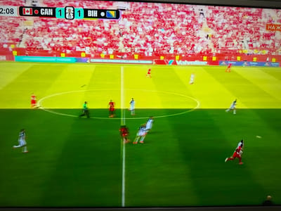 The image is a screenshot from a soccer match being broadcast on television. In the top left corner, a timer displays "2:08". To the right of the timer, a scoreboard indicates the teams "CAN" and "BIH" are tied 1-1. The background shows a stadium filled with a crowd dressed primarily in red. The field is green with yellow markings. Several players in white and red uniforms are visible on the field, some in motion as if participating in the game. A portion of a sign in Cyrillic script is visible on the right side of the screen. The photograph was taken of a television screen, as evidenced by the pixelation and glare. The provided location of Targovishte, Bulgaria is not supported by any visual information in the image.