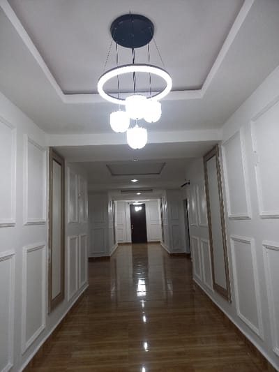 The image depicts a well-lit, empty indoor hallway, likely within a building in Oturkpo, Nigeria. The hallway features a glossy brown tiled floor with visible reflections of overhead lighting. Both side walls are uniformly paneled in white with decorative molding, creating a repeating pattern of rectangular sections. Several doors are integrated into these paneled walls, blending with the white decor; a darker, presumably wooden door is prominently visible at the far end of the corridor, marking its termination. The ceiling is white, multi-tiered with recessed lighting, and a prominent modern chandelier hangs centrally in the foreground. This chandelier consists of a large, illuminated circular LED ring with four smaller, spherical white light fixtures suspended beneath it. No people or specific actions are visible within the scene.