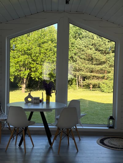 The image depicts a sunlit dining area inside a white-paneled room, with a large, triangular window offering a view of a lush green lawn and trees. A round, light-colored table with a dark, angular base stands in the foreground, set with three white chairs with wooden legs. On the table are decorative items including a white vase with a circular cutout, a rectangular holder with cutlery, a candle, and a yellow vase filled with purple flowers. To the right of the window, a black lantern sits on the floor next to a circular rug. The exterior scene shows a well-maintained lawn bordered by mature trees, suggesting a tranquil, possibly rural or suburban setting. There are no people present in the image. The lighting indicates it is daytime, with sunlight casting shadows on the floor and illuminating the foliage outside. The location is identified as Aleshkino DP, Russia.