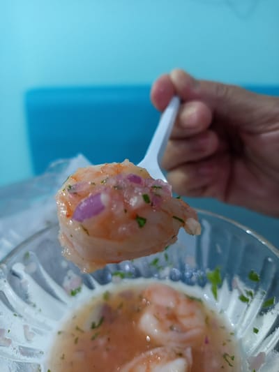 An extreme close-up shot shows a white plastic spoon lifting a piece of ceviche from a clear glass bowl. The ceviche appears to be made of shrimp, chopped red onions, and herbs, in a light pinkish liquid. The spoon is held by a hand, partially visible at the top right of the frame. In the background, a bright blue surface is visible. The scene is likely in Tolú, Colombia, a coastal town known for its fresh seafood and ceviche.