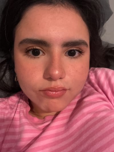A close-up, frontal medium shot captures a young person's face, with dark hair framing their features. They are wearing a pink and white striped t-shirt. The person's skin appears flushed, with subtle speckles of freckles visible. Their eyebrows are dark and full, and their eyelashes are long and dark. The eyes are open and dark, reflecting light. The nose is slightly shiny, and the lips are glossy, with a slight pout. The background is a dark, indistinct surface. The scene is set in Austin, Texas, United States of America.