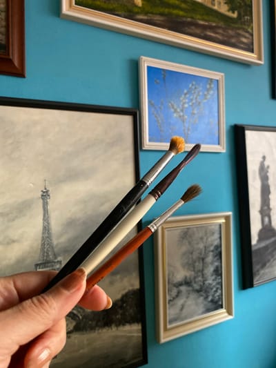 A person's hand, with light-toned skin and manicured nails, holds three paintbrushes of varying sizes and colors, positioned in front of a blue wall adorned with framed artworks. The paintbrushes, from left to right, have black, white, and reddish-brown handles. The leftmost brush has a fluffy, orange-tipped head. Behind the hand and brushes, on the wall, are several framed pictures. One prominent artwork on the left is a black and white depiction of the Eiffel Tower. To its right, a smaller framed picture shows a blue sky with white flowers. Further to the right, another framed artwork features a grayscale image of a statue. A larger framed picture with a light-colored frame is positioned below the blue sky artwork, showing a grayscale landscape. The overall scene appears to be an artist's workspace or a collection of artwork displayed on a wall. The location in Bílina, Czechia is not discernible from the image.