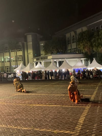 The image shows a night scene in front of a building in Kecamatan Batu, Indonesia, located at latitude -7.867 and longitude 112.514. There are numerous white tents in the foreground, lined with spectators. Two people are in the foreground, facing away from the camera, dressed in animal costumes, kneeling on the brick pavement. The pavement appears to be a parking lot or walkway. The scene is brightly lit. The building behind the tents is a two-story structure with lit windows. Palm trees are visible in the distance.