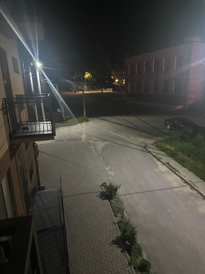 An outdoor, night-time, high-angle view captures a street scene with buildings and a paved walkway in Strășeni, Moldova. The foreground features a partial view of a building with a metal-railed balcony and a paved walkway with a brick pattern. To the right, a road is visible with tire marks and a dark-colored car parked along the side. A building with multiple windows, appearing to be a two-story structure, is situated across the street. The lighting suggests artificial illumination from streetlights, casting long shadows across the scene. Vegetation, including bushes and grass, lines the edge of the road. In the distance, a few indistinct figures can be seen walking on the street. The overall atmosphere is one of a quiet urban environment at night.