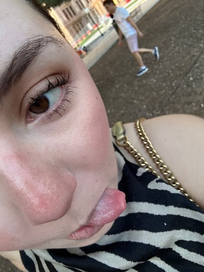 A selfie is tilted, focusing on the face with an extended tongue. The left eye is open, showcasing dark lashes and a brown iris. A double gold chain necklace rests on the bare shoulder. A black and white zebra-striped garment is visible. In the distant background, a person wearing white and pink clothing is on the ground in the bottom left corner. The background is generally blurry.