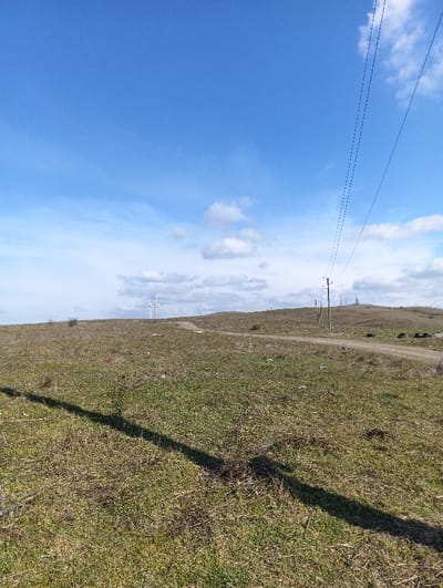 The image displays an open, rural landscape in Krumovo Gradishte, Bulgaria, under a sky with both clear blue areas and scattered white-grey clouds. The foreground consists of dry, patchy green and brown grass with some sparse vegetation and incidental debris. A prominent dark shadow extends diagonally from the lower left towards the center of the frame. In the midground, an unpaved dirt road curves from the right side, ascending a gentle slope into the distance. Parallel to the road on the right, a series of wooden utility poles support multiple overhead power lines. The background reveals rolling hills, with two white wind turbines visible on the left horizon and additional utility structures faintly discernible on the right. No individuals or active subjects are present in the scene.